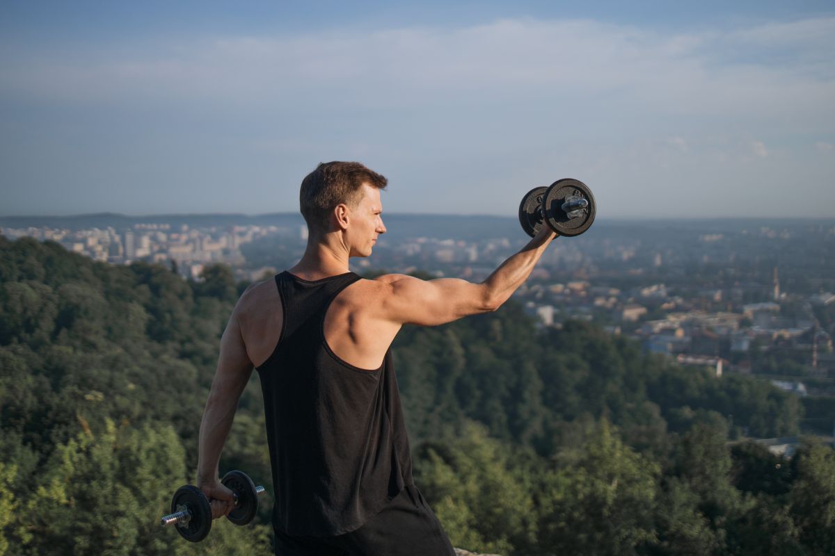 Muscular young man in activewear training arms with dumbbells outdoors. Caucasian male athlete having active workout on fresh air. Motivation concept.