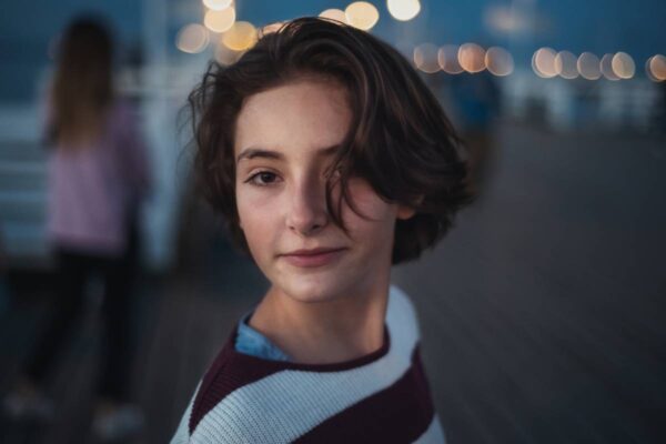 A portrait of preteen girl looking at camera outdoors on pier by sea at dusk, holiday concept.