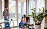 Young pretty couple in home clothes and slippers sitting at kitchen with large windows talking together about new project while drinking morning coffee and smartphone waiting for their friend to come.