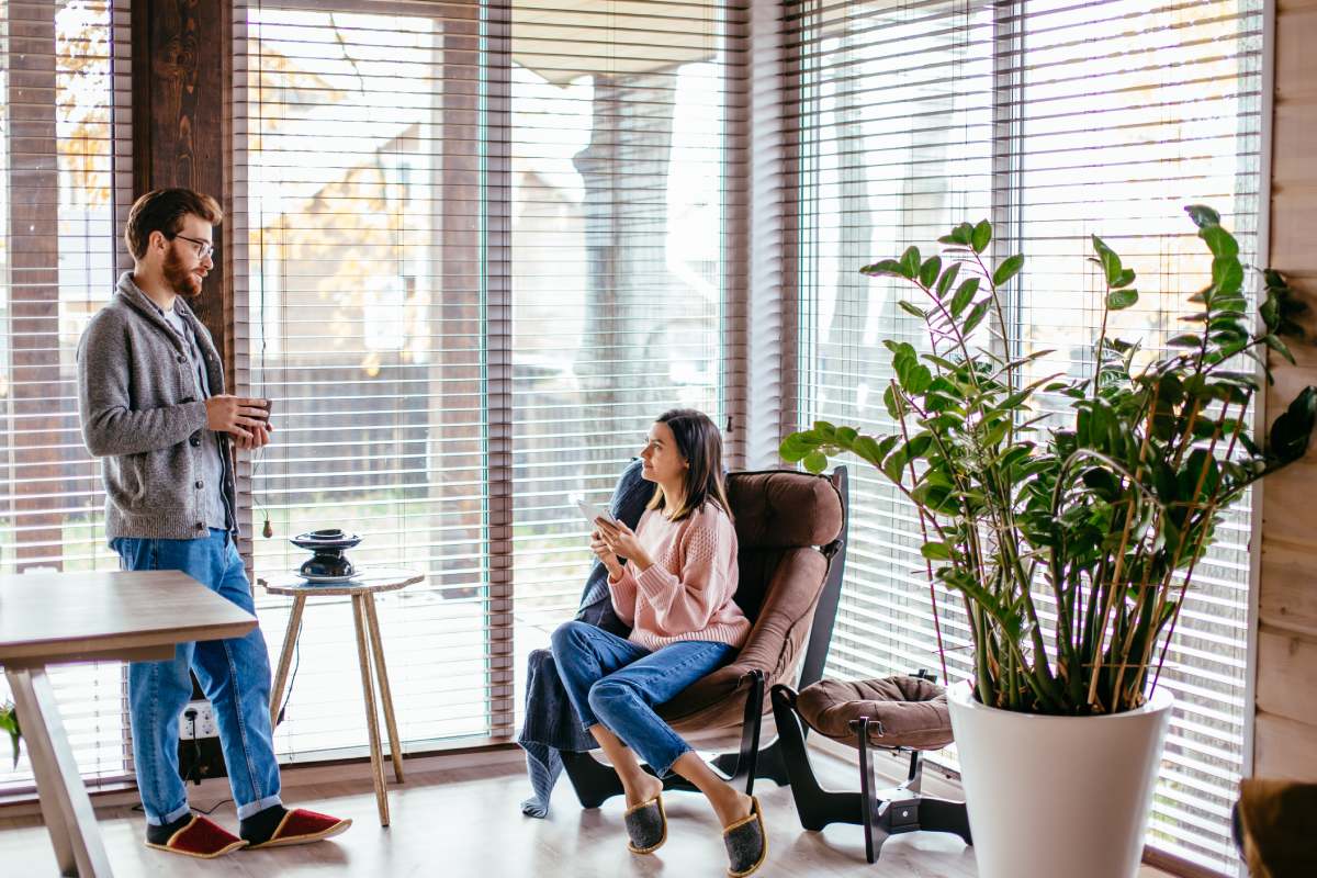 Young pretty couple in home clothes and slippers sitting at kitchen with large windows talking together about new project while drinking morning coffee and smartphone waiting for their friend to come.