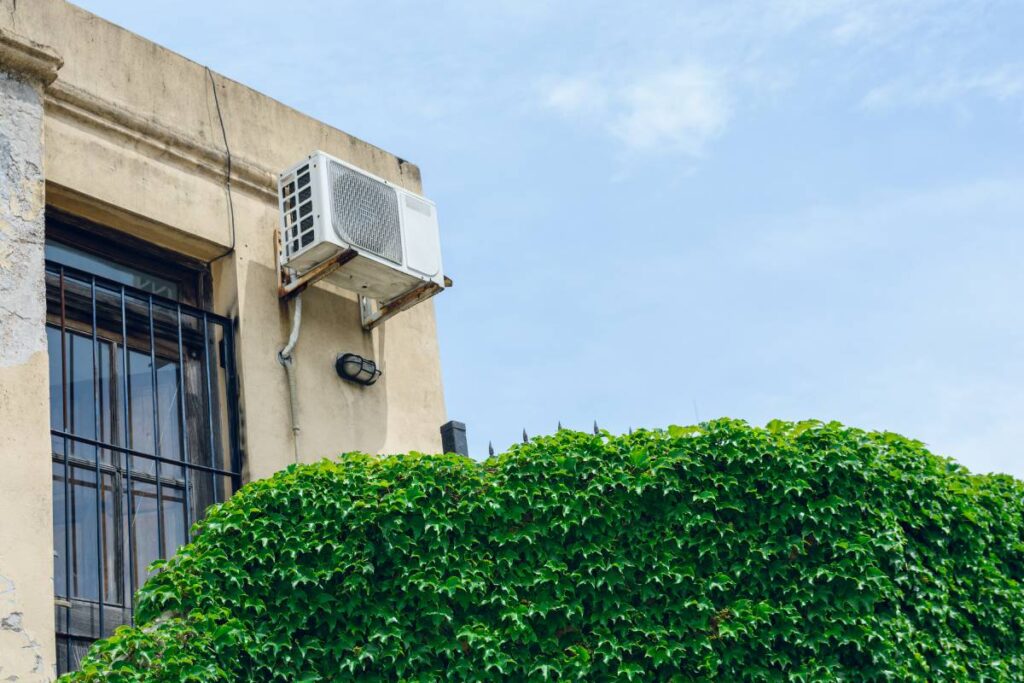 outdoor air conditioner unit in old house with tangled plant wall in foreground and clear blue sky in background, concept of conservation and climate change, copy space